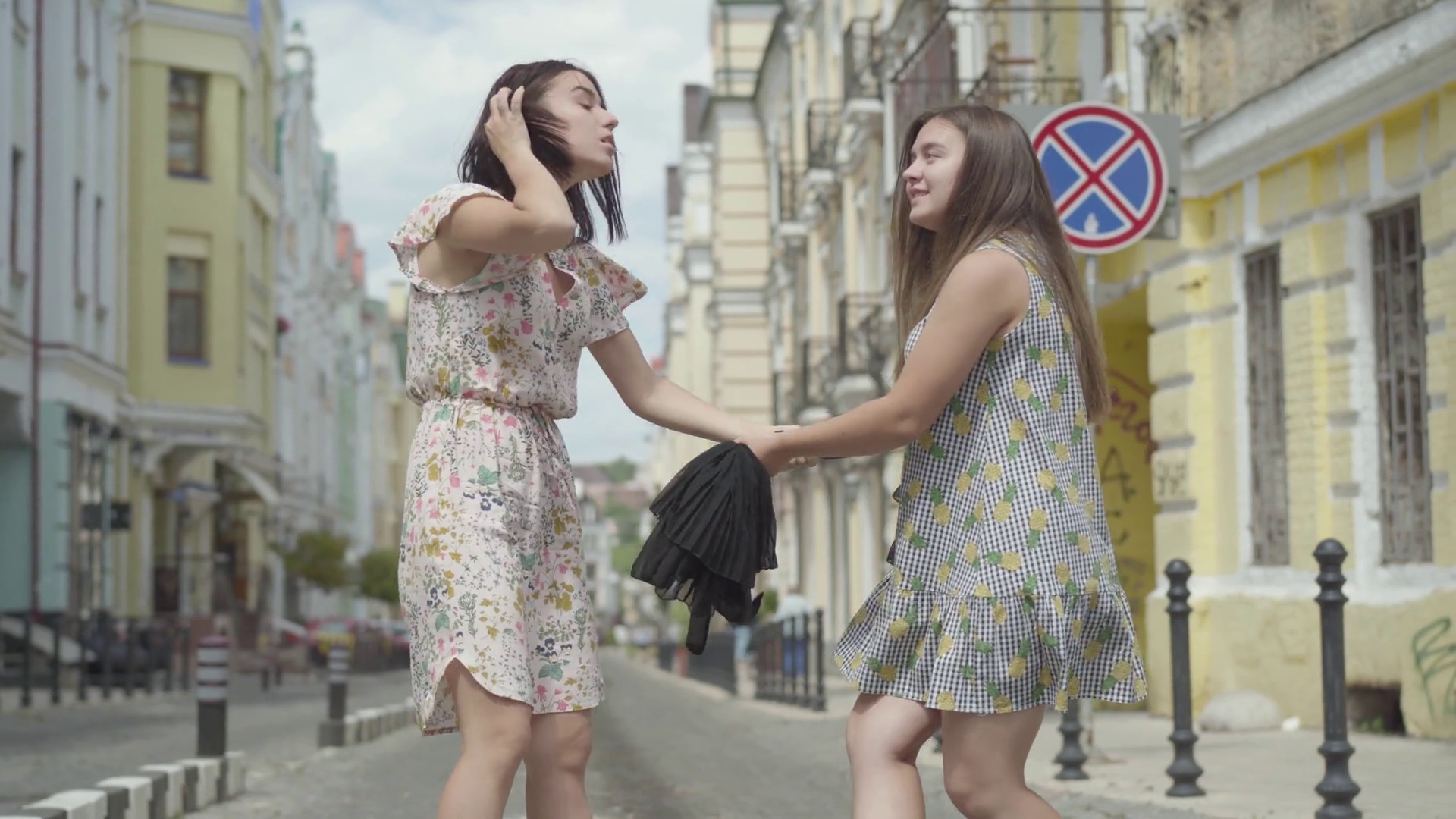 Two fashionable girls holding the black dress and pulling it to their sides  on the street. Shopping concept. Fashionistas are fighting for stylish  clothes