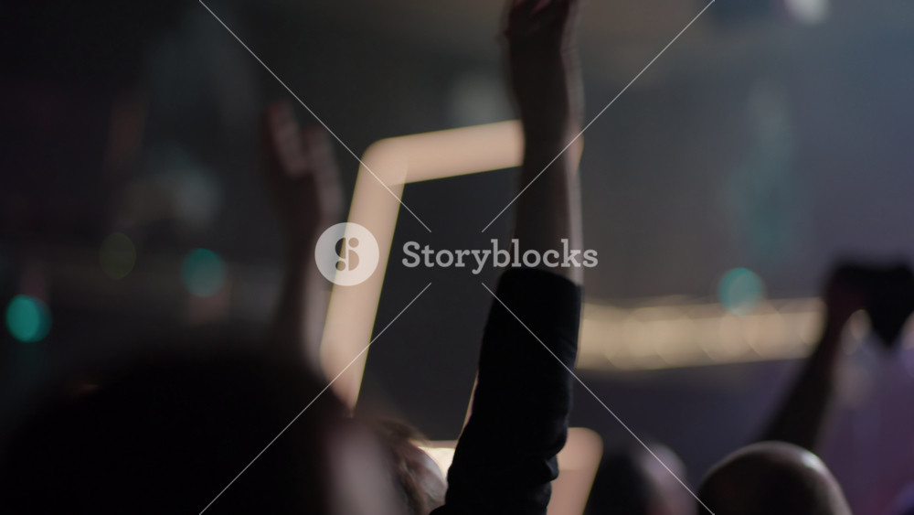 Blurred shot of energetic crowd at music festival. People wave hands in air in front of glowing stage lights. Silhouettes of fans cheering