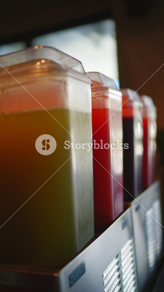 A slow, cinematic pan shot across a row of colorful fruit juices in beverage dispensers. Close-up with shallow depth of field highlights