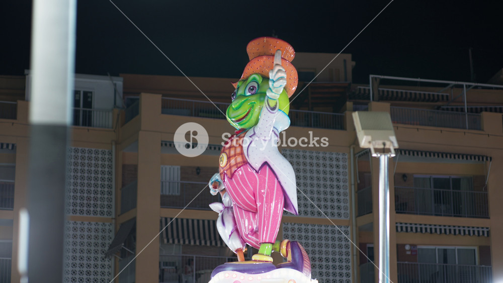 Colorful papier mache festival figure of whimsical cartoon character in top hat, mounted on rotating monument at night, residential