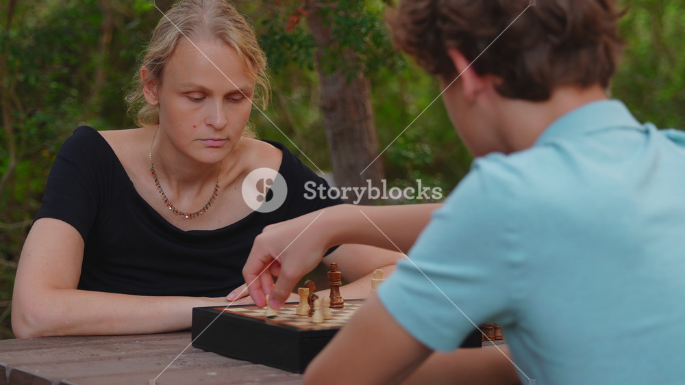 A mother and her teenage son play a game of chess on a wooden table in a park, focusing on the board as they take turns moving their chess