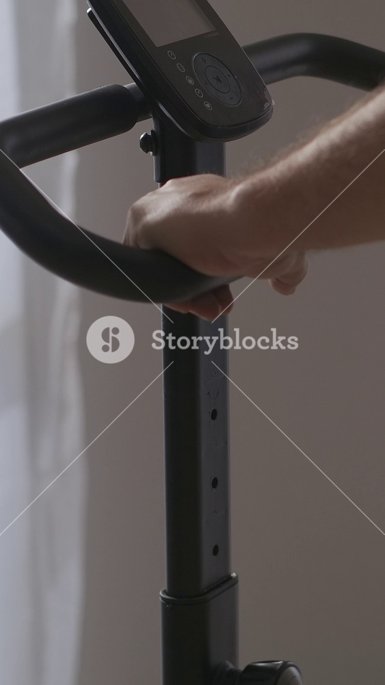 Close-up of a person gripping the handlebar of an exercise bike with a digital display screen in a softly lit room showing details of