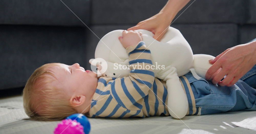 Adorable baby lying on the floor wearing a striped shirt with blue pants interacting with an adult pouring milk from a bottle, showcasing a
