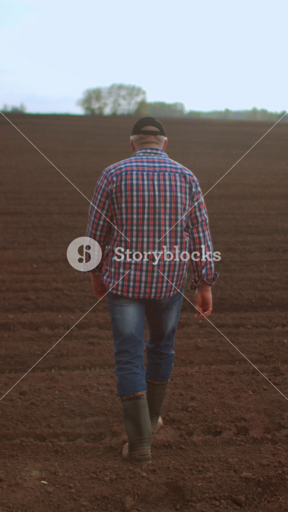 Solitary farmer walking across freshly tilled brown field in rural countryside landscape, wearing plaid shirt, jeans and boots under soft