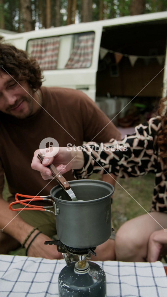 Vertical shot of young woman in casualwear sitting next to her boyfriend and mixing soup or other food in pan on electric appliance while cooking something for breakfast or lunch