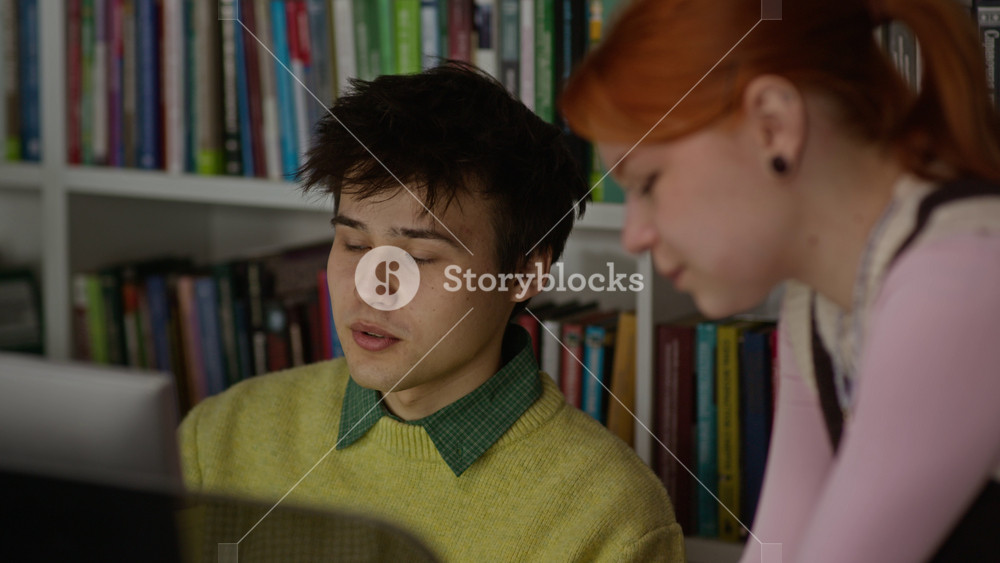 Close up on Caucasian teenage boy and Caucasian teenage girl studying together in library, looking at computer screen and discussing project. Footage of academic collaboration for e-learning ads