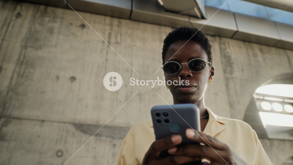 African American young adult woman wearing sunglasses standing near concrete wall, using smartphone next to blue suitcase. Video is perfect for travel apps, mobile booking, and tourism marketing