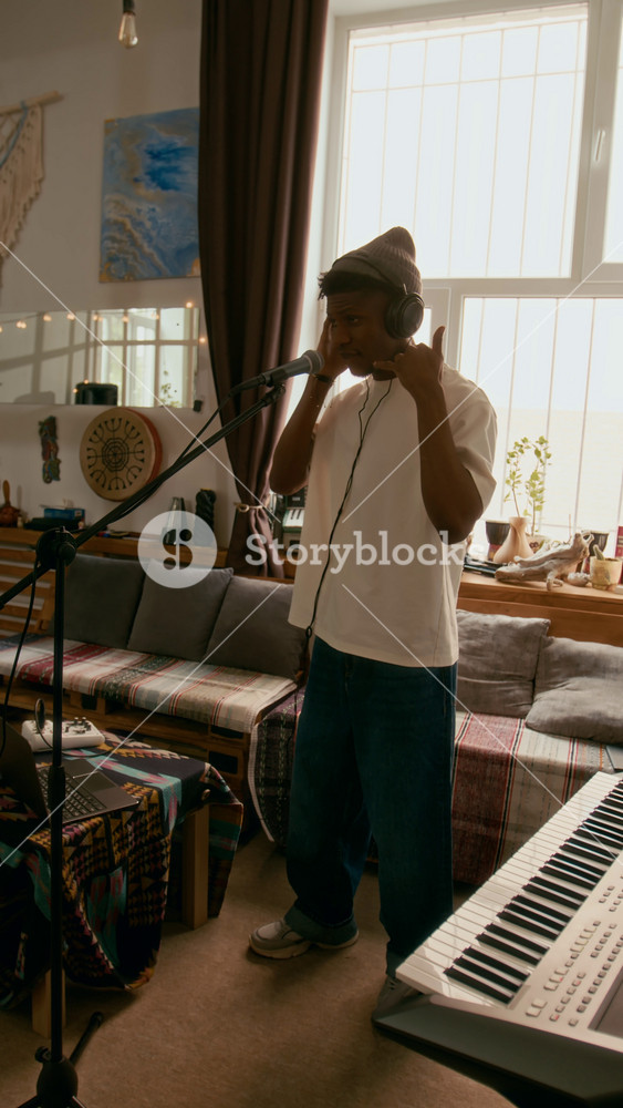 Vertical shot of young African American musician in casual attire and headphones standing in front of microphone in home studio and performing his new song during repetition