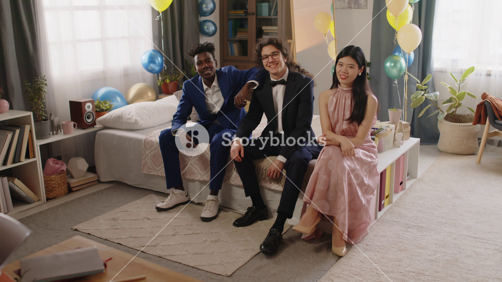 Happy teenage guys and girl in tuxedos and evening gown sitting on double bed in domestic room decorated with balloons and looking at camera while waiting for prom party