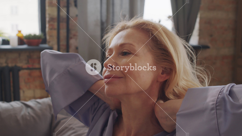 Handheld medium close-up portrait of senior Caucasian woman with blond hair wearing comfortable lavender silk pyjamas rubbing her neck and fixing hair