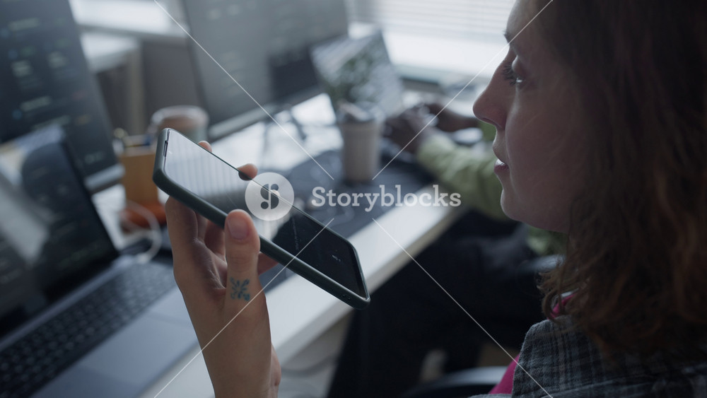 Close up shot of young adult Caucasian woman holding smartphone and recording voice command for AI assistant in office while colleague works in background. For mobile app and voice messaging marketing