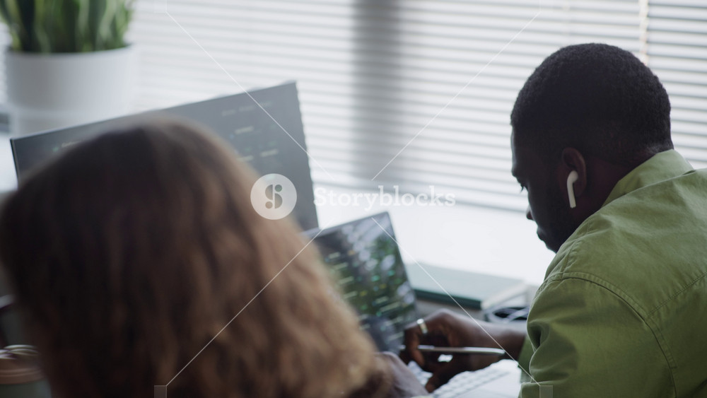 Side view of African American young adult man and Caucasian young adult woman working on computers in office, female developer putting on headphones. Footage ideal for tech startup recruitment videos