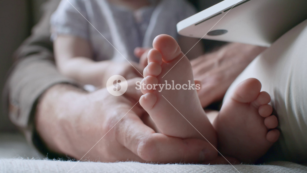 Handheld close up shot of hands of unrecognizable father holding little feet of toddler girl sitting beside him and watching cartoons on laptop