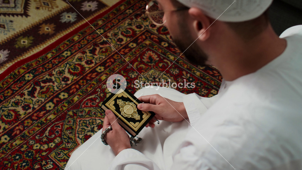 High angle of young man in white skullcap and thobe sitting on his knees on carpet, closing Quran after reading it, kissing holy book and performing prayer in the morning