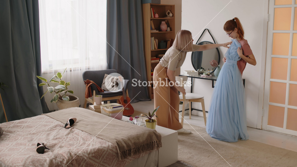 Mature mother standing in front of her teenage daughter and helping her try on long blue fatin evening dress while both preparing for prom celebration before graduation event