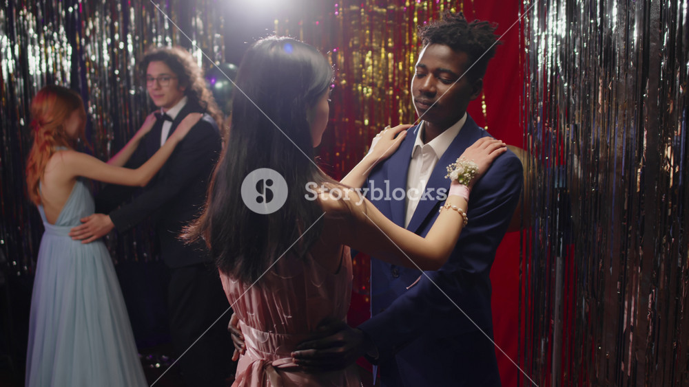 Two intercultural teenage couples in classy attire dancing in front of camera in the night club at prom party while guys keeping hands on waists of their girlfriends