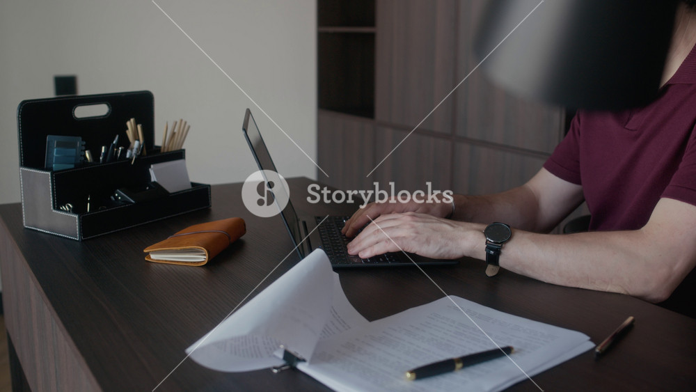 Medium close up of hands of mature male writer sitting at desk in home office typing on laptop, printed draft and pen placed nearby on desk