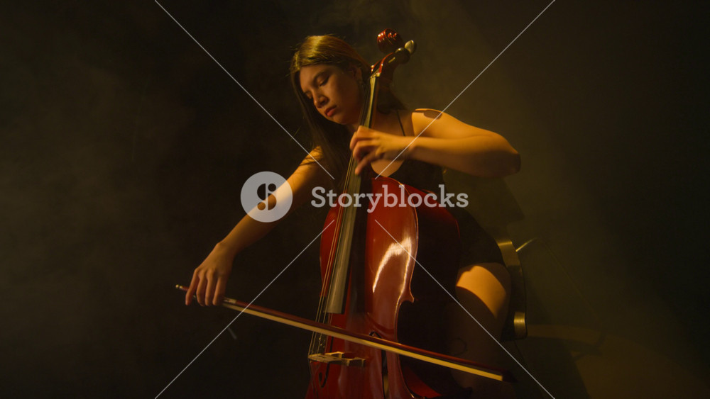Concentrated young adult Hispanic female musician wearing black evening dress playing cello while performing acoustic music on fog filled stage under warm spotlight, slow motion