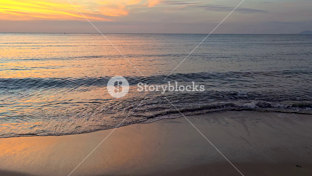 A serene view of the calm ocean waves gently lapping against the sandy shore in Chumphon, Thailand. The vibrant colors of the sunset fill
