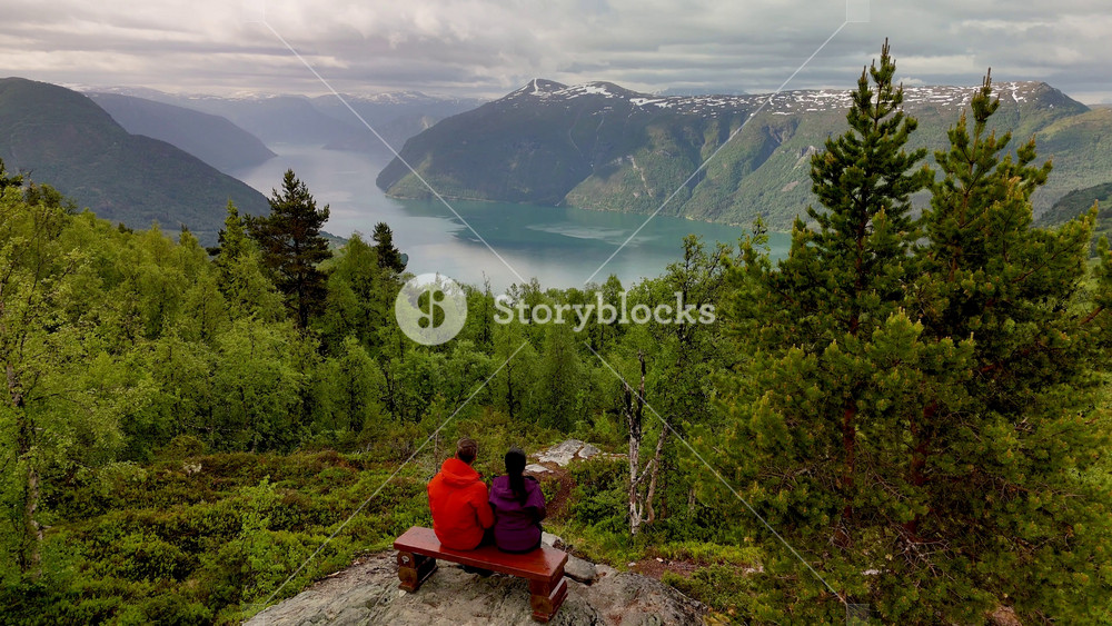 Two individuals relax on a rocky outcrop, taking in breathtaking views of a serene fjord surrounded by lush greenery and majestic mountains