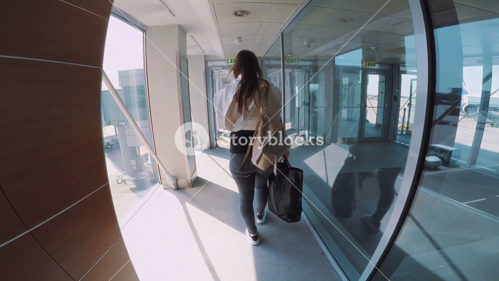 Female traveler walking through airport corridor towards airplane gate, carrying a black bag, with reflections on glass walls and bright sunlight illuminating the pathway