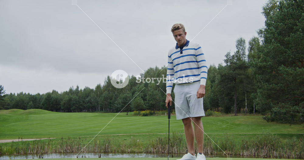 A young stylishly dressed man in sportswear takes aim and hits a ball with a golf club, trying to drive it into the hole, but fails and the