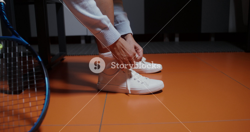 Woman tennis player tying shoelaces on white sneakers while sitting on a bench in a locker room, about to play tennis, legs close-up