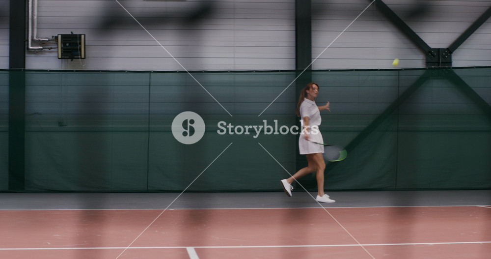 A young woman tennis player, dressed in a white sports uniform, plays tennis, hitting the ball over and over again, actively moving. Filming