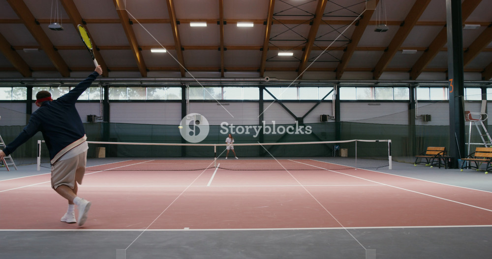 Young man and woman play tennis on an indoor tennis court, hitting blow after blow with a racket, moving actively. Video filming from behind