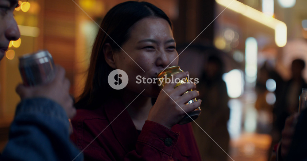A woman drinks beer from a tin can and talks to her friends. They are standing on a city street in the evening. Close-up of her face