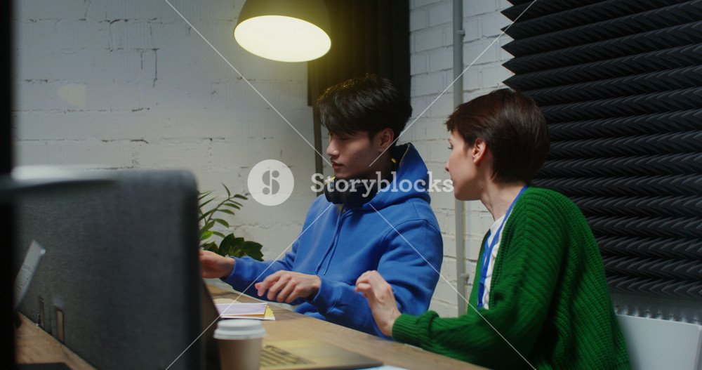 A young beautiful woman of European appearance and an Asian man in headphones, talking during the working day, sitting at the same table and
