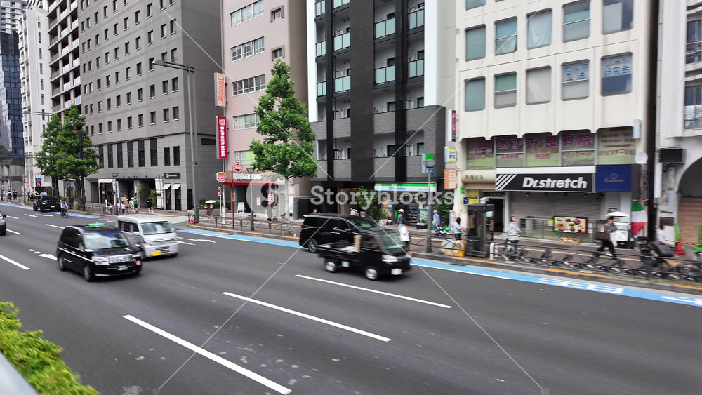 TOKYO - May 24, 2026 - Forward three-quarter driving view of a busy downtown Tokyo street with shops, restaurants, vehicles, pedestrians, and modern urban architecture. 15190