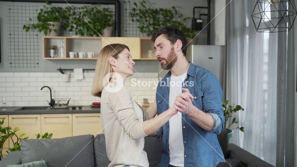 Happy adult man and woman dancing slow dance in daytime in kitchen at home. A cheerful married couple looks at each other, smiles, romantically circling in a dance. They have a good festive mood