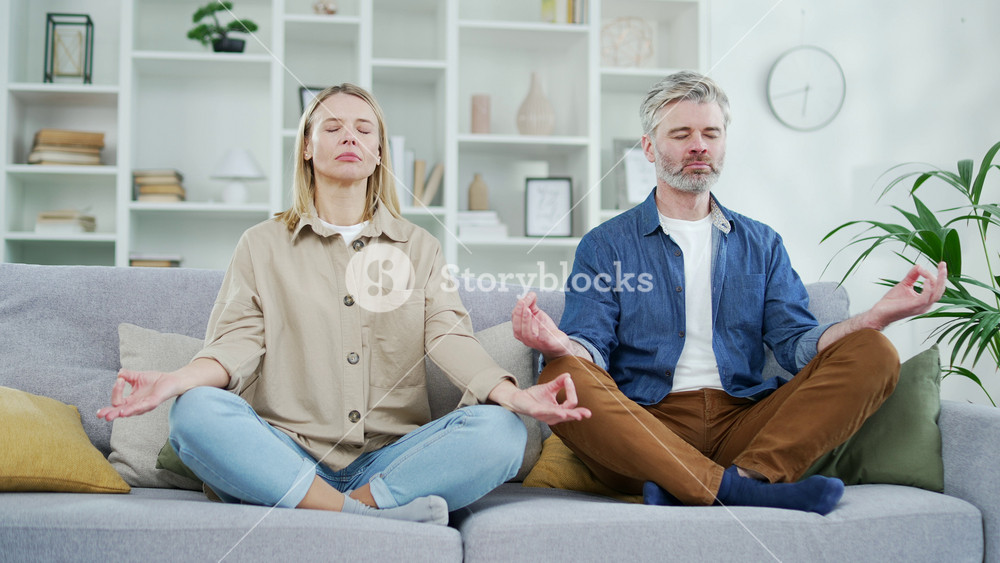 Mature married couple meditating with eyes closed in lotus position while sitting on sofa in living room at home. Husband and wife practice yoga, breathing calmly on a comfortable couch