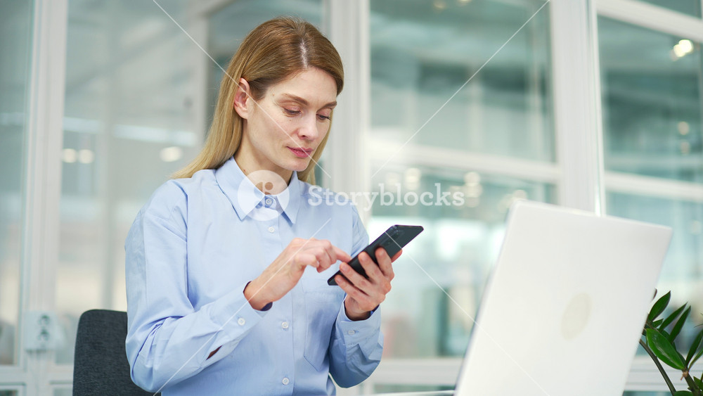 Happy excited businesswoman read great news on smartphone while sitting at workplace in modern office. A smiling and satisfied female employee is joyfully reviewing a pleasant message notification