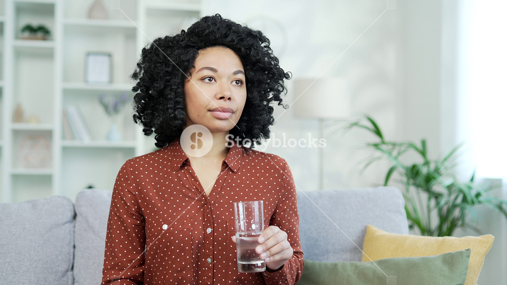 A young african american female drinks water from a glass while sitting on the sofa in living room at home Happy smiling black woman feeling relieved, enjoying a clean cool drink, relaxing and resting