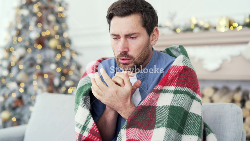 Upset bearded man covered with a blanket froze sitting on sofa in living room at home during winter New Year Christmas Xmas holidays. Male cold indoors. Guy feels discomfort due to low temperature