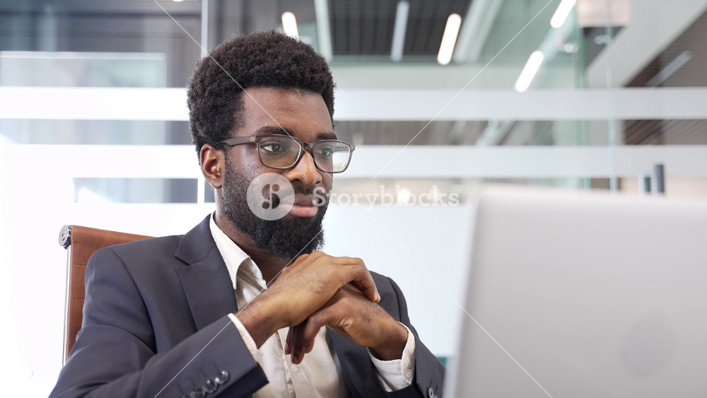 Happy handsome african american businessman in formal suit receiving good news on laptop sitting at desk in business office. Satisfied bearded entrepreneur expressing happiness and success. Close up