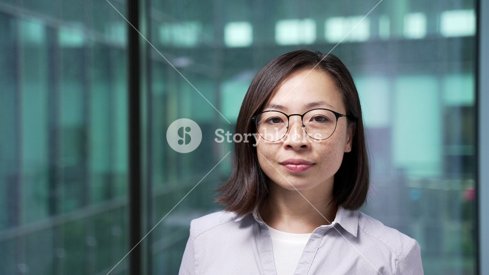Portrait of serious asian businesswoman standing at workplace in modern glass business office. Head shot of confident female manager or employee in glasses and casual shirt looking at camera. Close up