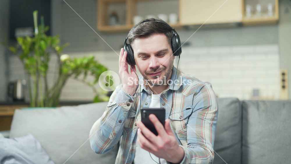 happy young bearded cheerful handsome caucasian man enjoying listening to music sitting at home on sofa in room with headphones using smartphone. The male dances and sings Cheerful rhythmic joyful guy