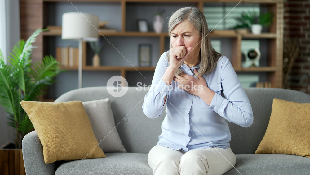 Sick senior gray haired female coughing sitting on sofa in living room at home. Sick elderly retired woman with a cold or flu feels bad, holds her hand to her chest. She has fever or bronchitis