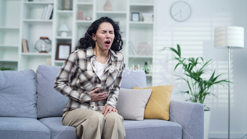 Young adult brunette woman suffering from abdominal pain while sitting on sofa in living room at home. Sad female keeps her hands on her stomach She has poisoning, constipation, spasms or menstruation