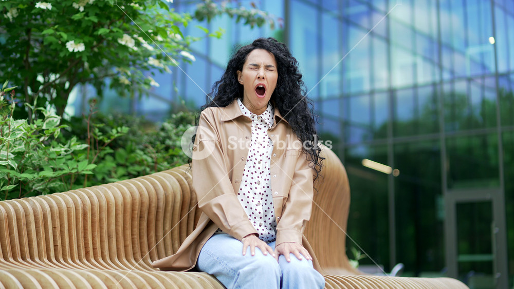 Overworked young female employee feels tired and wants to sleep while sitting on a bench on the street near an office building. Sleepy exhausted brunette woman yawns, closes her eyes. Overload at work