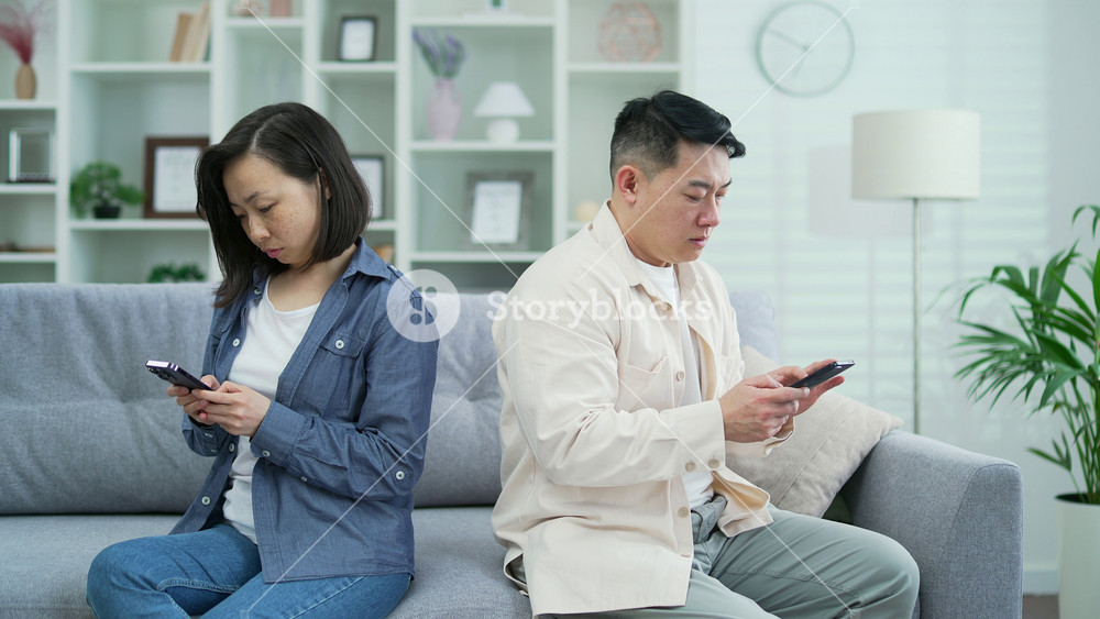 Asian wife and husband, offended at each other, look at their smartphones while sitting side by side on the sofa in the room at home. Conflict in a married couple. Problem, crisis in family relations