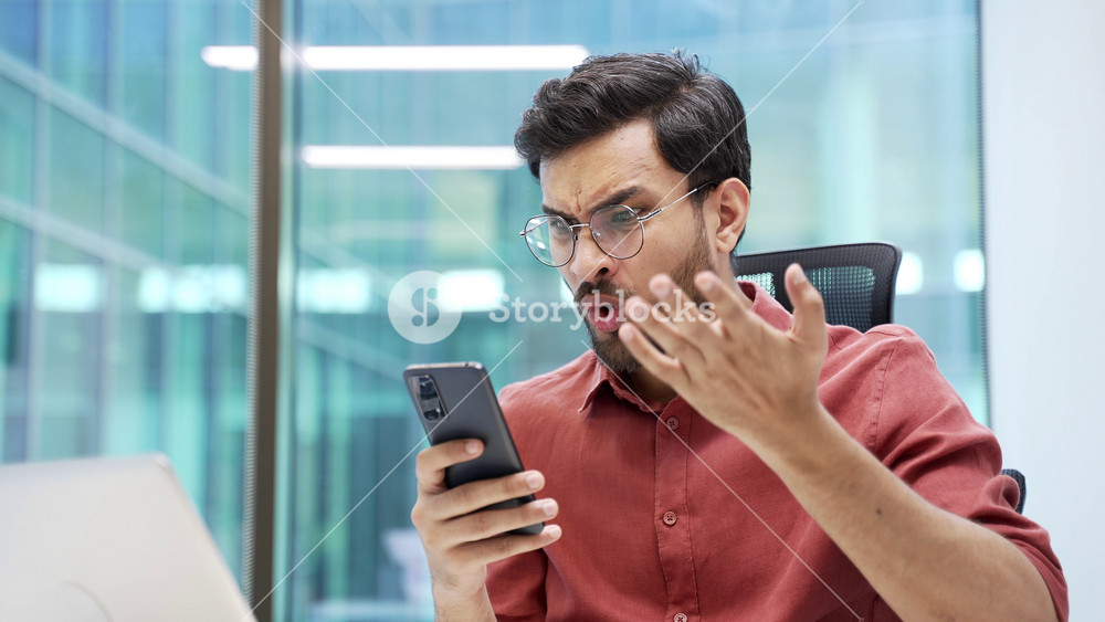 Shocked stressed businessman reads bad news on smartphone while sitting at desk at workplace in business office. Frustrated worried worker is sad reviewing negative information on a phone. Close up