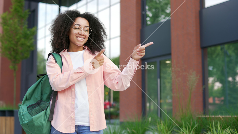 Portrait of smiling african american female student pointing fingers at empty copy space for writing text and looking at camera. Young woman standing near university building. Template for advertising