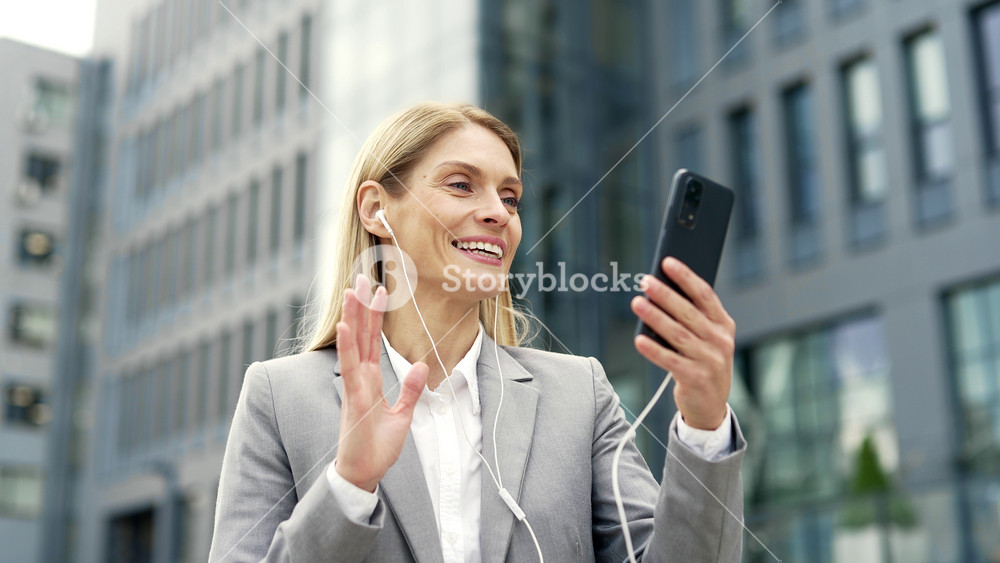 Smiling businesswoman talking on a video call using a phone and headphones while standing near an office building. Happy mature woman in formal suit has conversation with a client or business partner