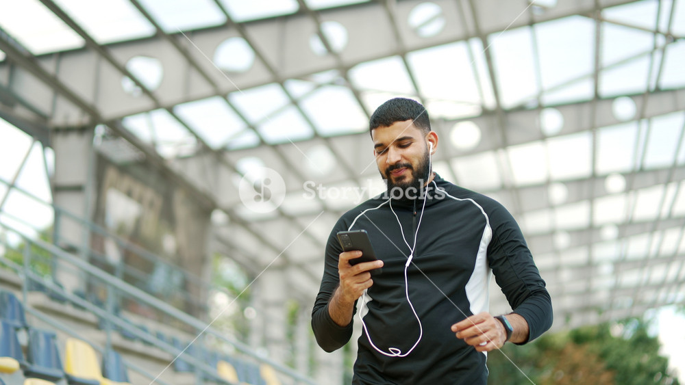 Adult bearded smiling sportsman walking with headphones and mobile phone enjoying listening to music outdoors on urban city stadium. Happy joyful male athlete in sportswear relaxing after training