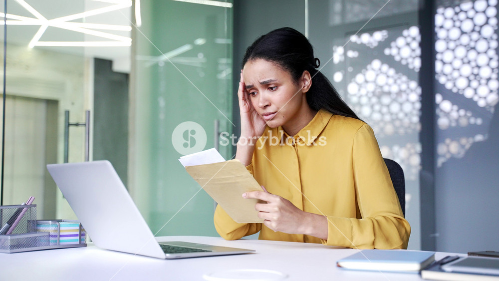 Sad young businesswoman opening envelope and reading letter with bad news at office desk. Disappointed female employee looking at document and feeling upset after receiving negative message.