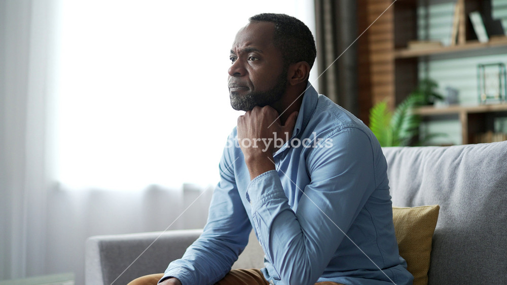 Silhouette of mature african american male feels depressed sitting on sofa in living room at home. Sad lonely senior black man thinking about his problems. He is unhappy, disappointed and has bad mood